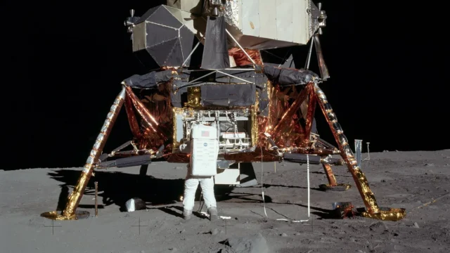 Astronaut in a white spacesuit stands next to the gold-foil-covered Apollo Lunar Module on the grey lunar surface under a