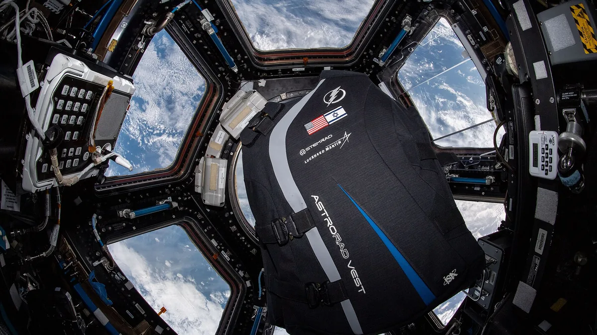 AstroRad vest with US and Israeli flags floats inside the ISS Cupola with Earth in the background.