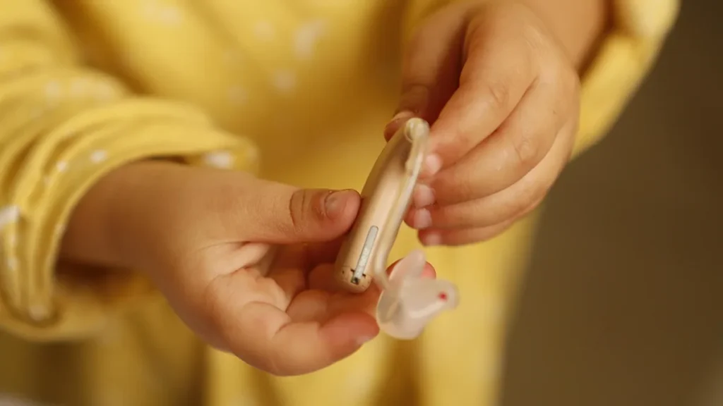 Child's hands holding a beige behind-the-ear hearing aid with a clear plastic earpiece.