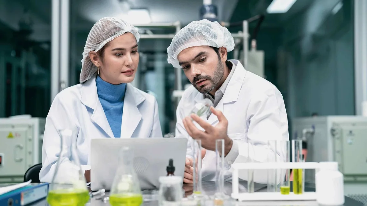 Two scientists in lab coats and hairnets examine a sample and laptop screen in a laboratory filled with test tubes.