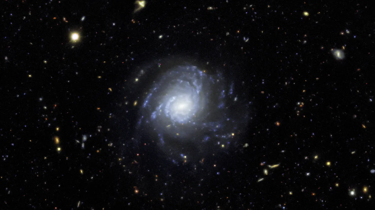 A bright spiral galaxy glows amidst a deep field of distant stars and smaller galaxies.