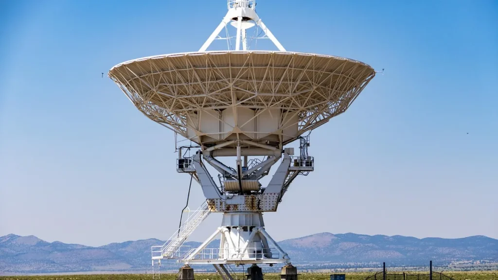A large white radio telescope dish stands against a blue sky with mountains in the background.