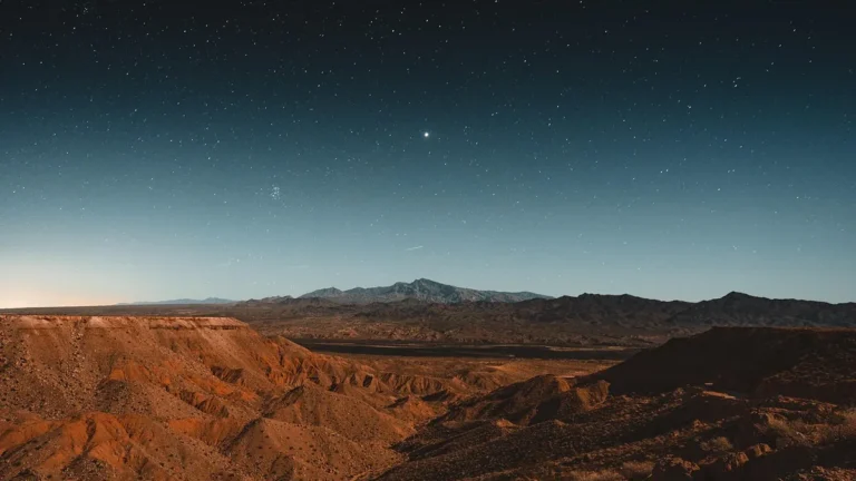 Starry night sky over a reddish desert mountain landscape with a bright star shining.