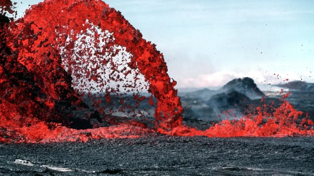Bright red molten lava erupts in a dramatic arch over a dark, cooled volcanic field.