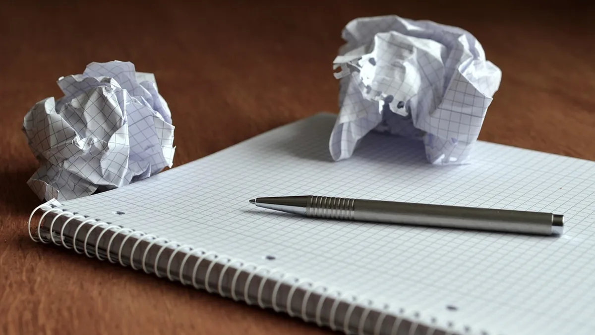 Silver pen on a grid spiral notebook with crumpled paper balls on a wooden table.