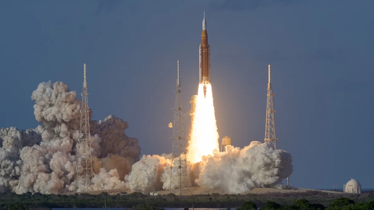 A massive rocket launches skyward, leaving giant plumes of white smoke and bright fire under a blue sky.