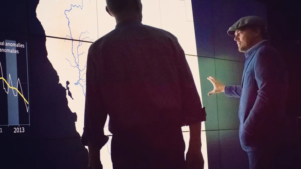 Leonardo DiCaprio gestures toward a climate anomaly map and graph displayed on a large bright screen.