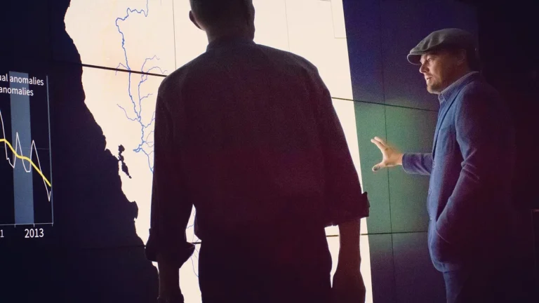 Leonardo DiCaprio gestures toward a climate anomaly map and graph displayed on a large bright screen.