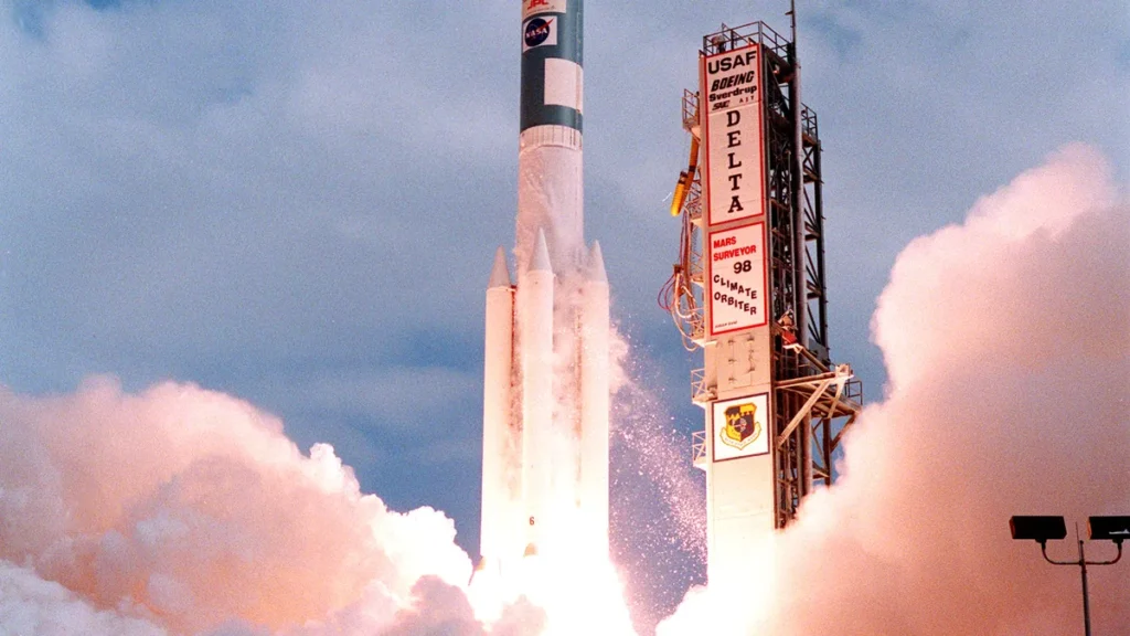 Delta II rocket launch with bright fire and billowing white clouds around the launch tower.