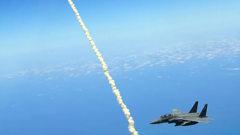 A military fighter jet flies alongside a thick, puffy white rocket trail high in the blue sky.