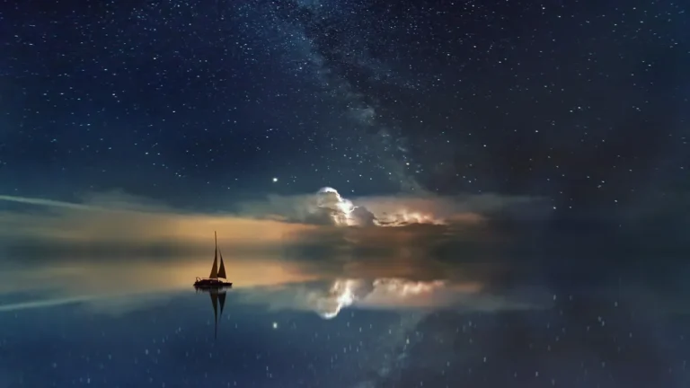 Silhouette of a sailboat on perfectly still water reflecting a star-filled sky with the Milky Way and glowing clouds.