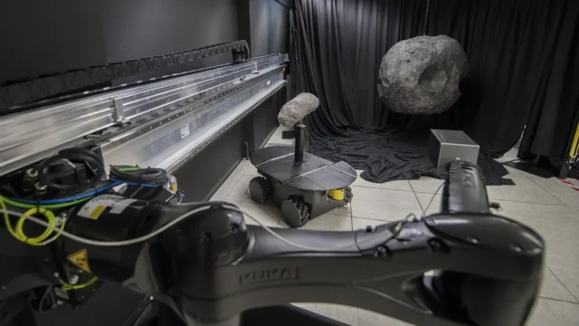 Close-up of a KUKA robotic arm in a lab with a small wheeled robot and a hanging mock asteroid.
