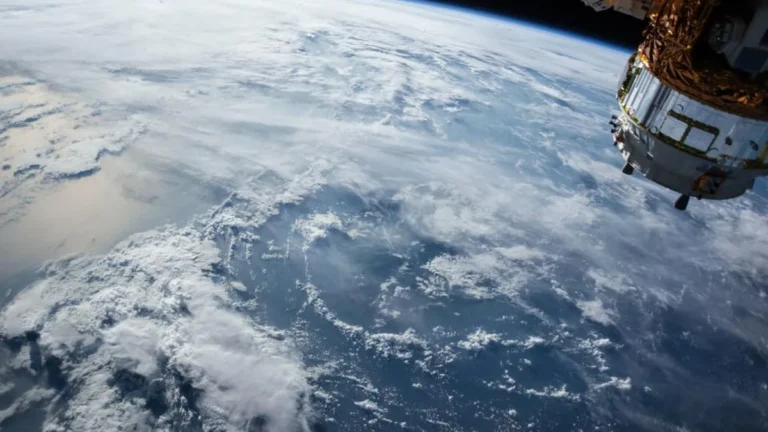 View of Earth's clouds and ocean from orbit, with part of a spacecraft visible in the upper right corner.