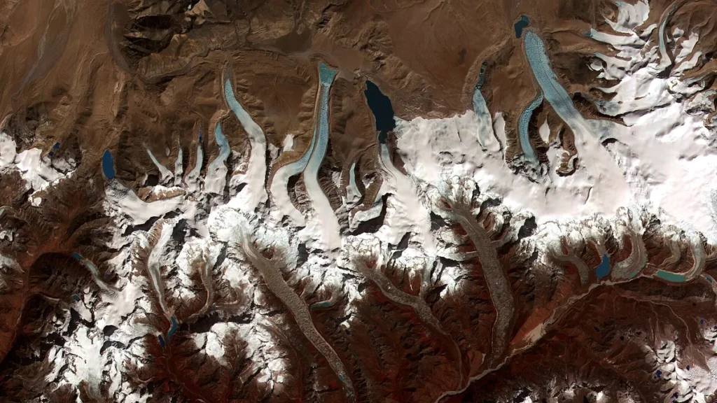 Satellite view of snow-capped mountains and winding blue glaciers stretching across a brown landscape.