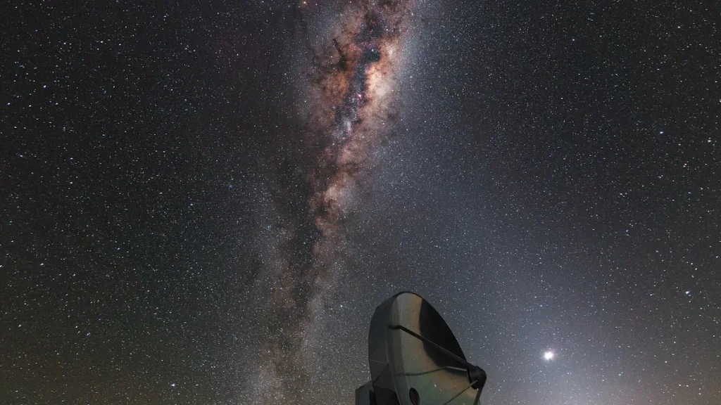 Milky Way arching over a radio telescope dish under a starry night sky with a bright celestial light.