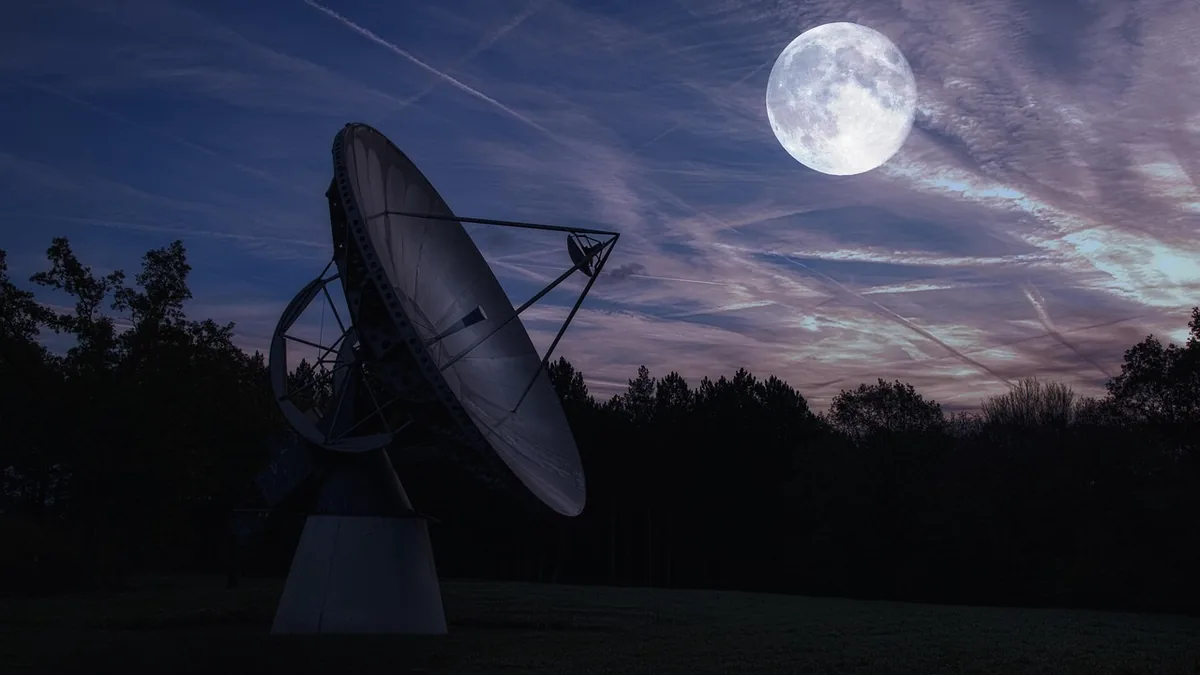 Large radio telescope silhouetted against a purple twilight sky with a bright full moon and wispy clouds.