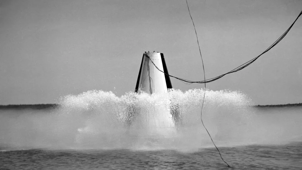 A black-and-white photo of a conical space capsule splashing down into water with a large spray.