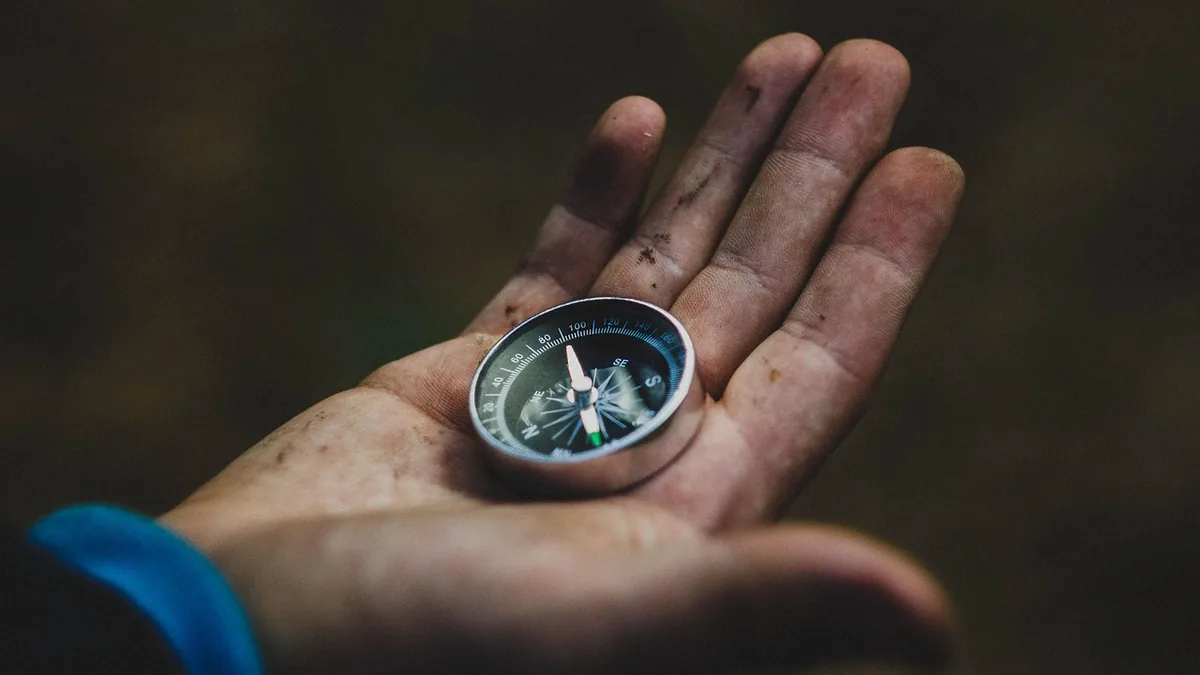 A close-up of a dirty hand holding a small silver compass, symbolizing magnetic navigation and orientation.