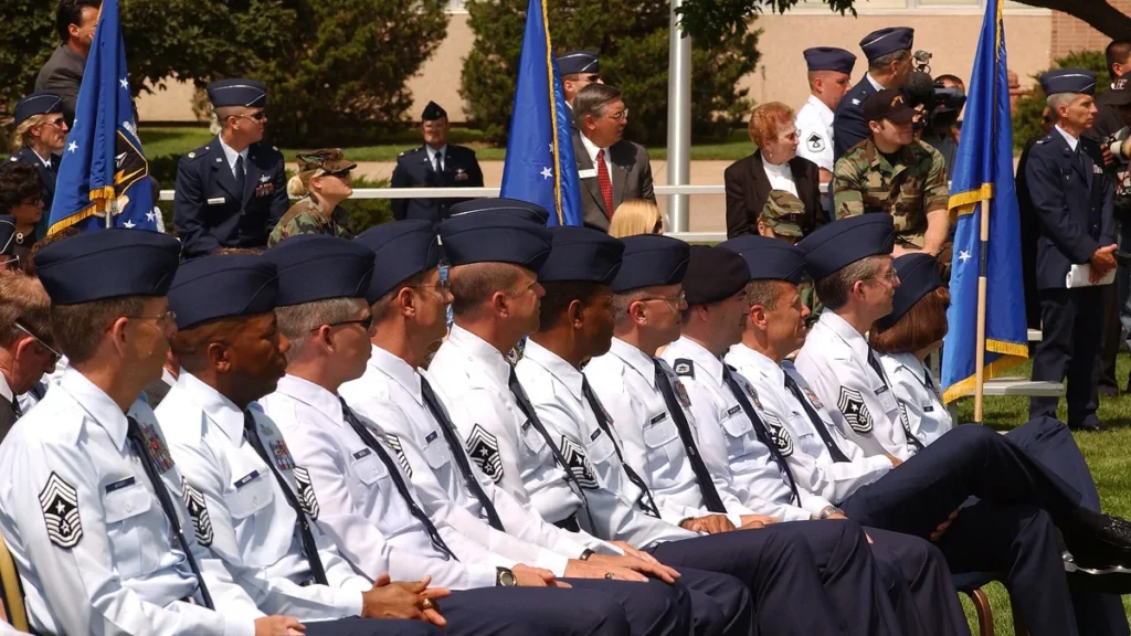 US Air Force chief master sergeants in white uniforms sit in a row during an outdoor ceremony.