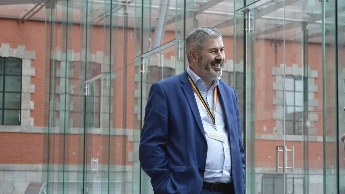 Smiling man in a blue suit and colorful lanyard stands before a glass wall with a red brick building.