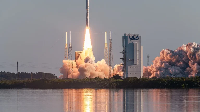 A ULA rocket launches into the sky, with bright fire and billowing smoke reflected in the foreground water.