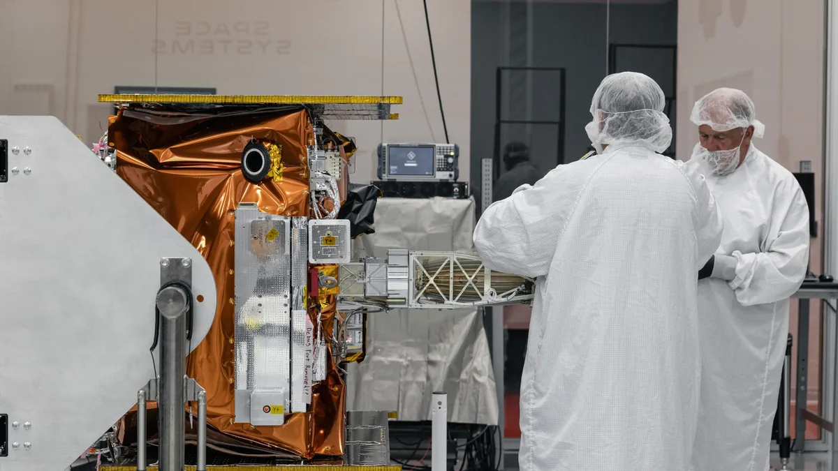 Technicians in cleanroom suits inspect a gold-foil-wrapped satellite component inside a cleanroom facility.