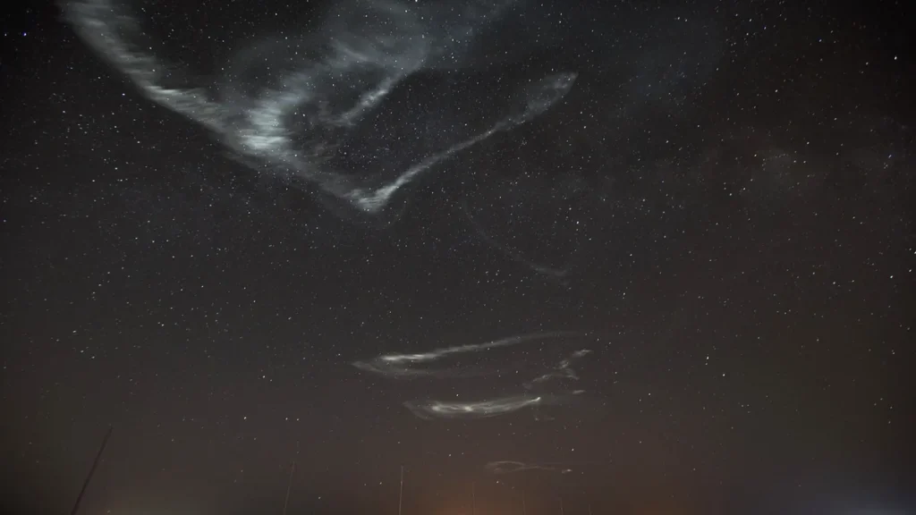 Wispy glowing rocket vapor trails drift across a brilliant starry night sky.