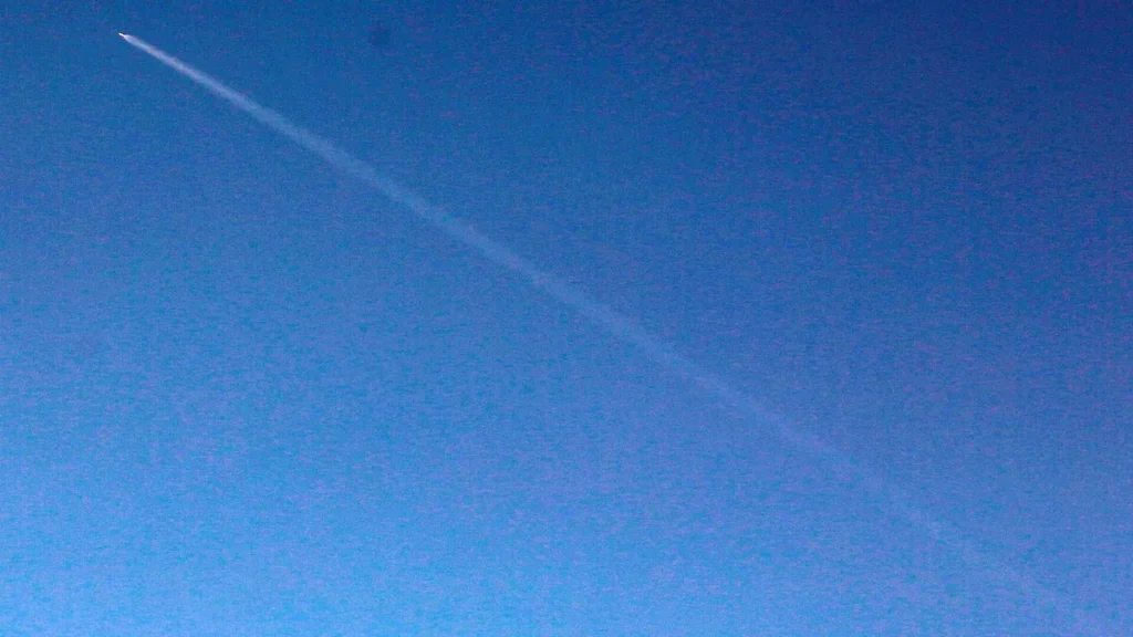 A bright rocket leaves a long white vapor trail streaking diagonally across a deep blue sky.