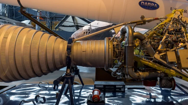 A large silver rocket engine on display, with a white rocket bearing the DARPA logo in the background.