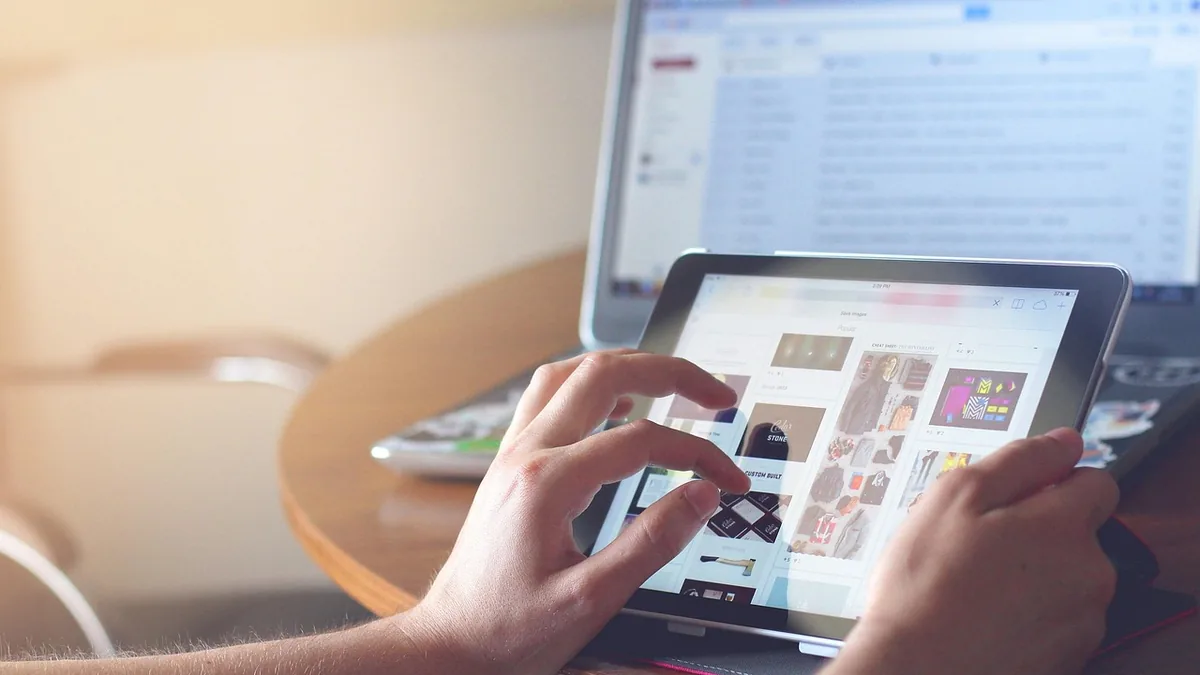 Close-up of hands holding and scrolling on a tablet displaying a grid of images, with a laptop in the background.