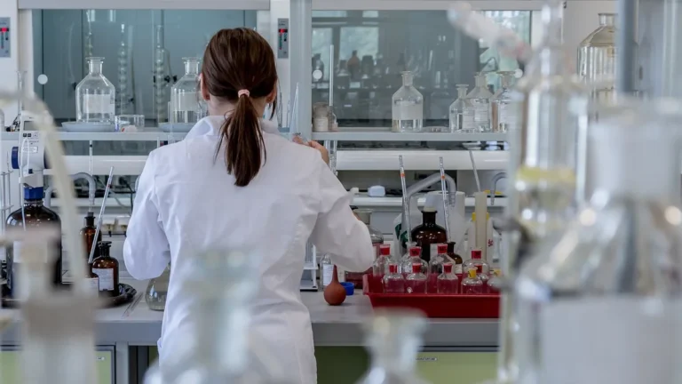A scientist in a white coat working in a laboratory filled with glassware and scientific equipment.