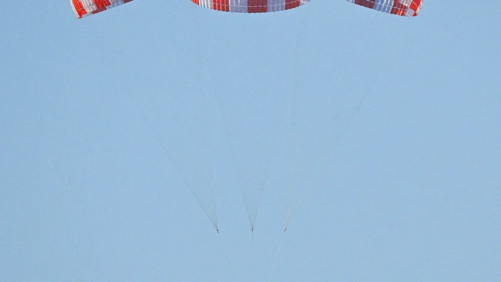 Red and white striped parachutes float against a clear blue sky, part of a space cargo recovery system test.