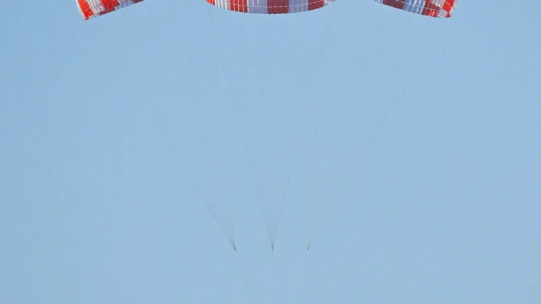 Red and white striped parachutes float against a clear blue sky, part of a space cargo recovery system test.