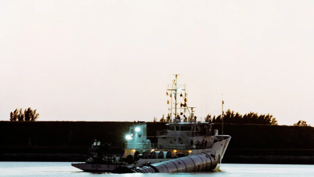 Large vessel towing a giant ribbed cylinder booster in water at dusk.