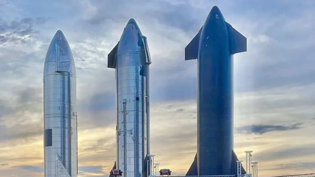 Three SpaceX Starship rockets stand against a cloudy sky, showing metallic and dark-colored hulls.