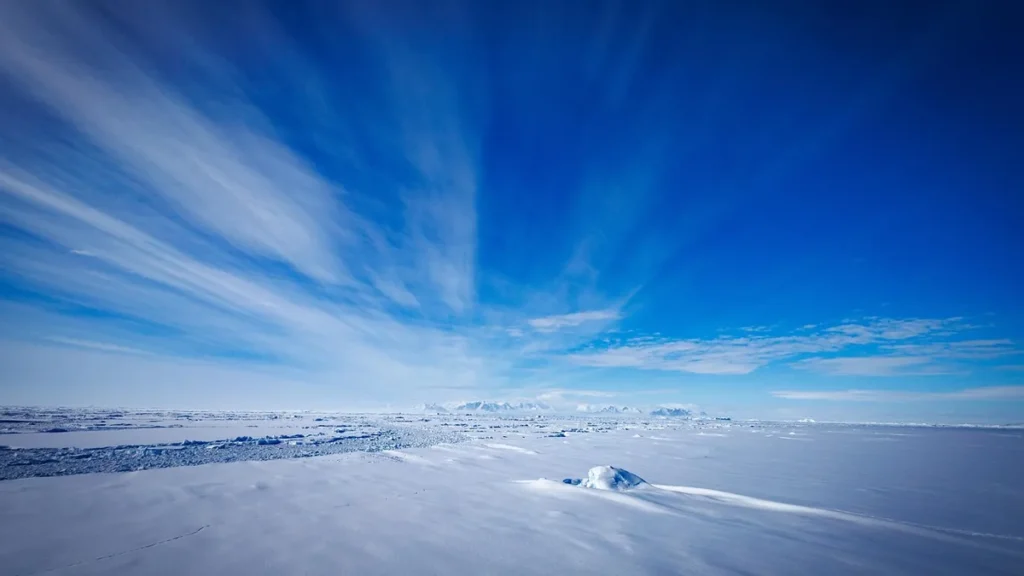Vast white ice sheet stretching to the horizon under a vibrant blue sky with wispy white clouds.