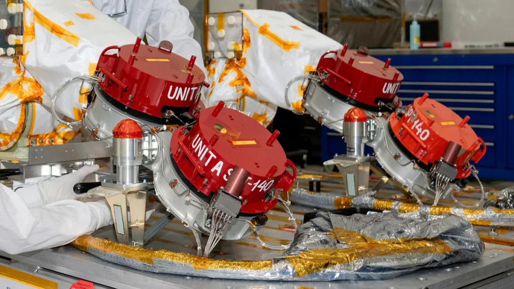 Red cylindrical spacecraft thrusters labeled UNITAS being assembled by technicians in a cleanroom.