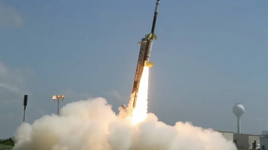A rocket launches into a blue sky, leaving a massive trail of bright white smoke and fire.