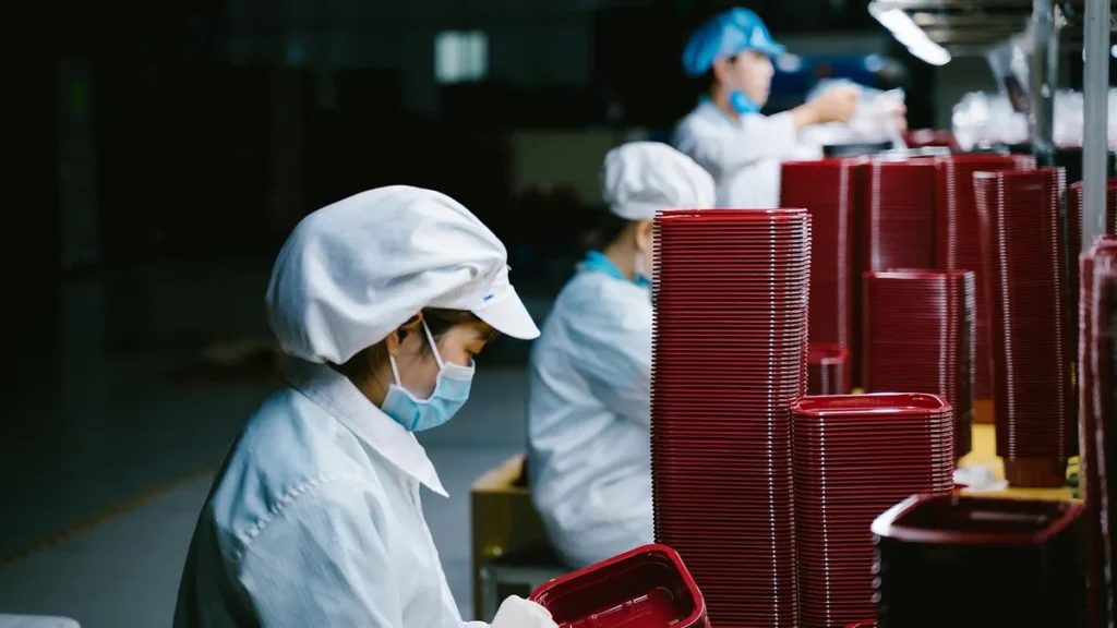 Workers in cleanroom suits stack red satellite components on an assembly line.