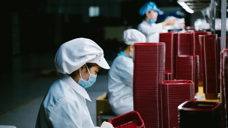 Workers in cleanroom suits stack red satellite components on an assembly line.
