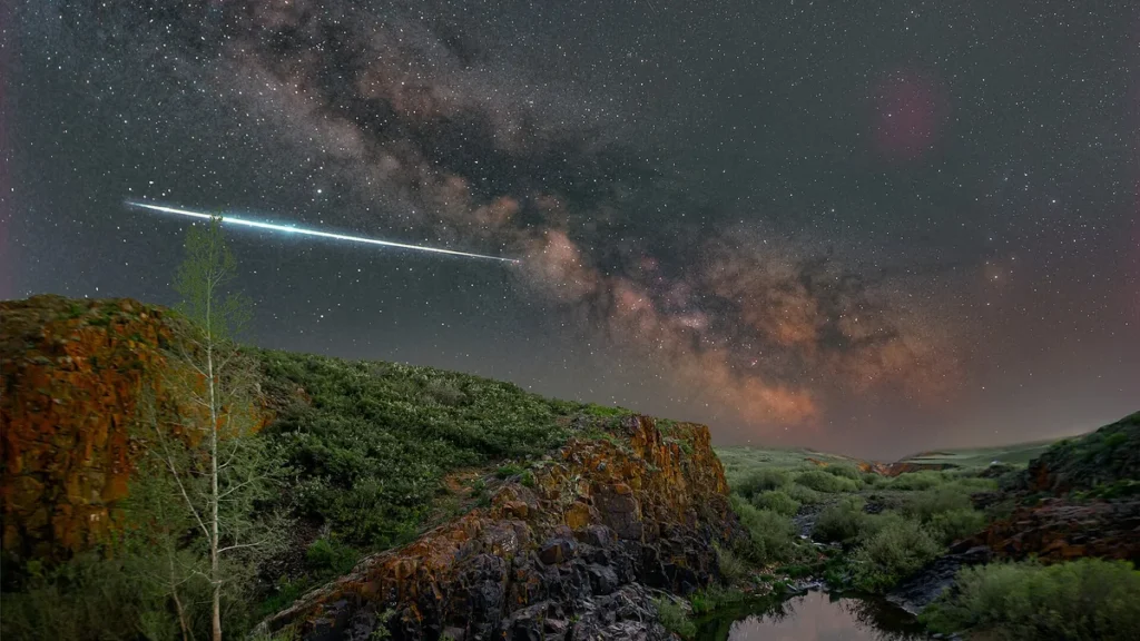 A bright meteor streaks across the Milky Way over a rocky green landscape under a starry sky.