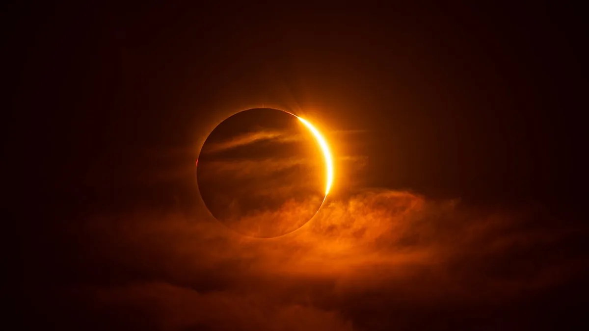 A solar eclipse shines with a bright orange crescent, partially veiled by dark clouds.