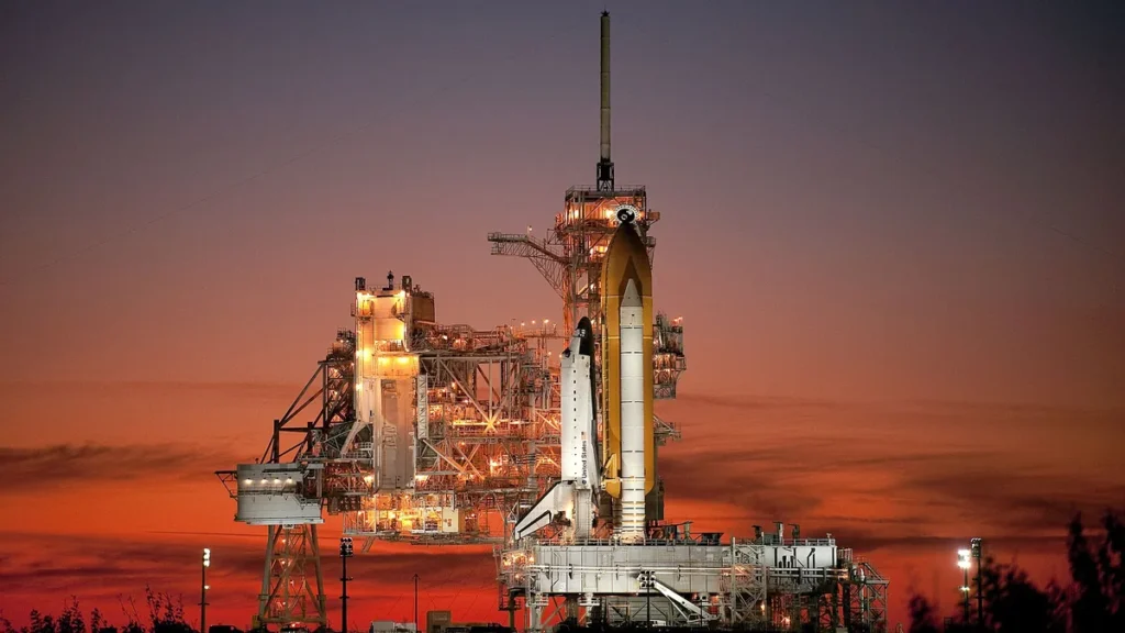 Space shuttle on launch pad illuminated against a vibrant orange sunset sky.
