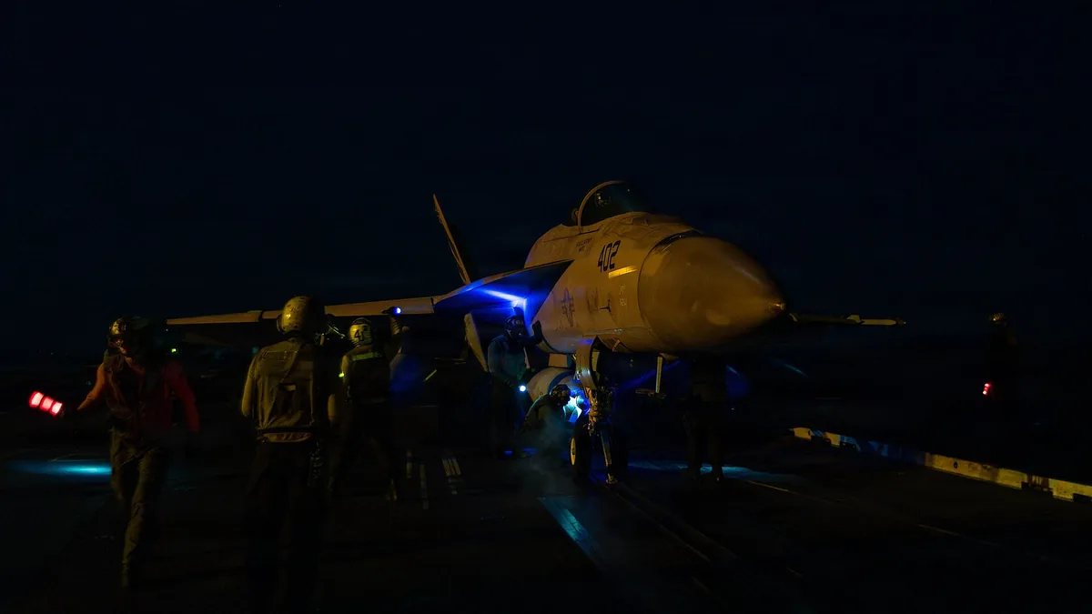 An F/A-18 jet on a dark flight deck surrounded by crew members holding blue and red signaling lights.