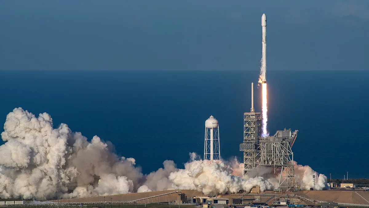 A SpaceX Falcon 9 rocket launches over the ocean, leaving a huge billowing cloud of white smoke.