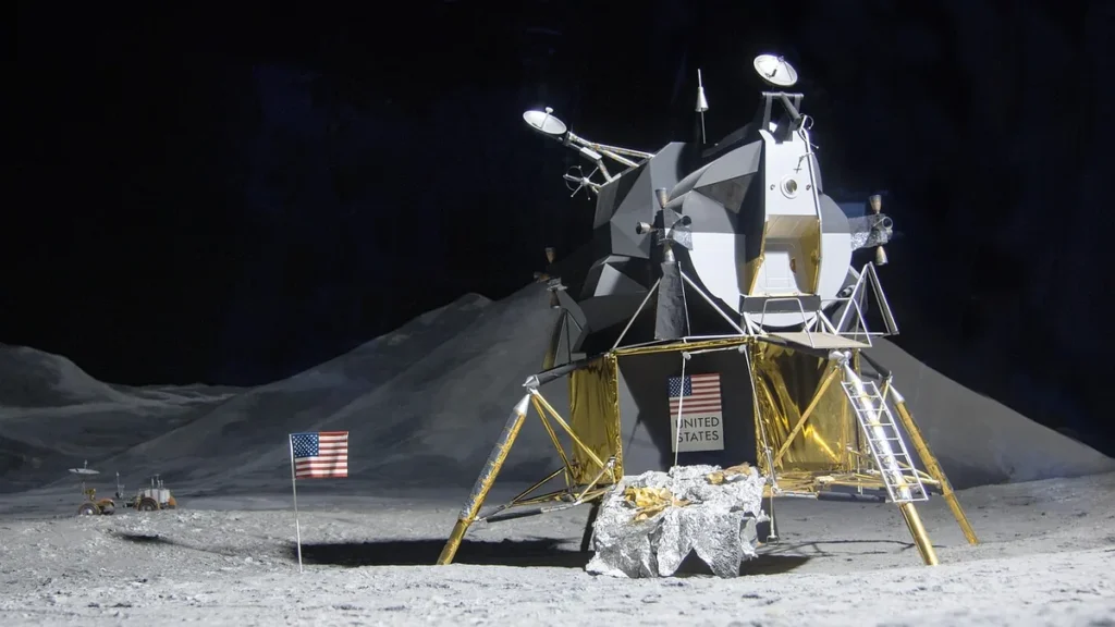 Model of an Apollo Lunar Module on a simulated lunar surface with a small US flag.