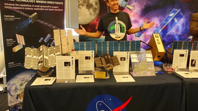 A presenter stands behind a NASA display table showcasing small spacecraft models and technology factsheets.