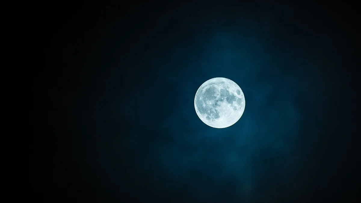 A luminous full moon shines brightly against a dark, blue-tinted cloudy night sky.