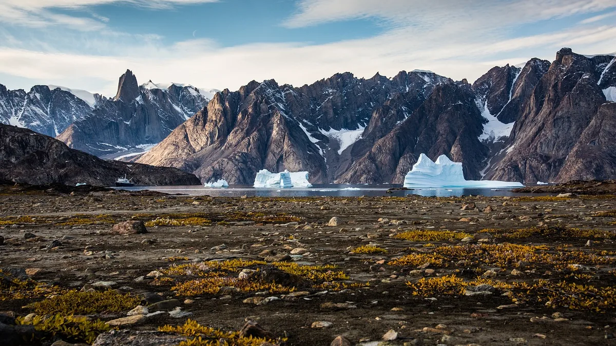 Arctic landscape with rocky tundra, icebergs floating in a fjord, and jagged snow-capped mountains in the background.