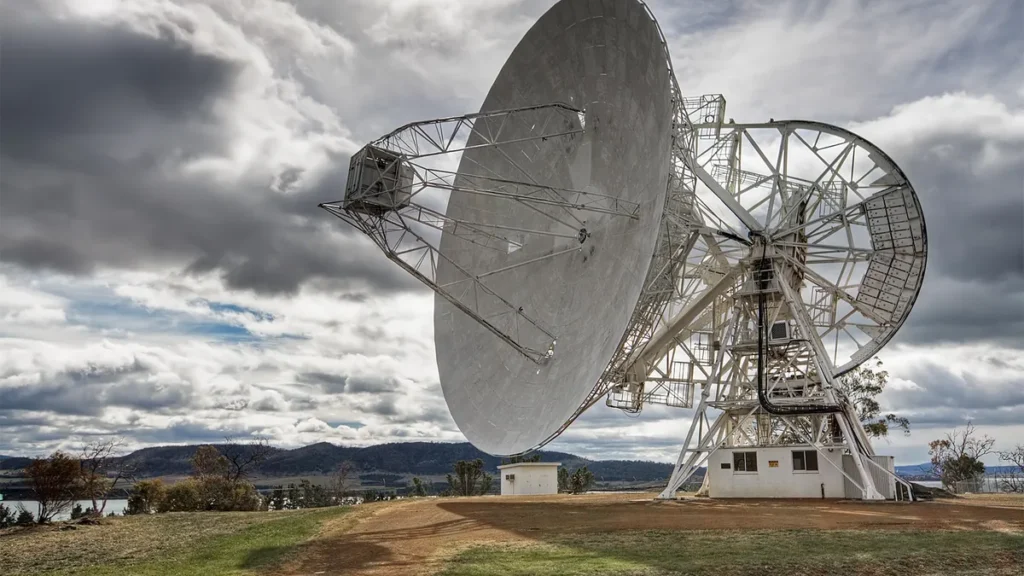 Large white satellite dish antenna standing against a dramatic cloudy sky, representing space communication infrastructure.
